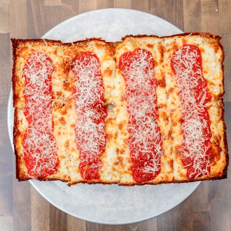Rectangular Detroit-style pizza with thick crust, red sauce stripes, and shredded cheese on a metal tray.
