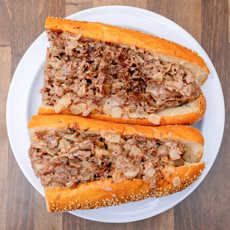 Two halves of a cheesesteak sandwich, filled with steak and onions, on a white plate.