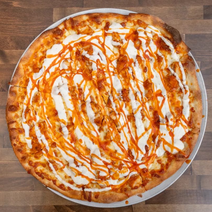 Overhead view of a buffalo chicken pizza with ranch and hot sauce drizzle on a wooden table.